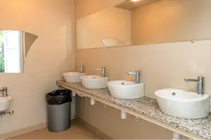 Mehrere Waschbecken in einem modernen Sanitärraum, mit Steinwaschtisch, Armaturen und Abfalleimer an der Wand.