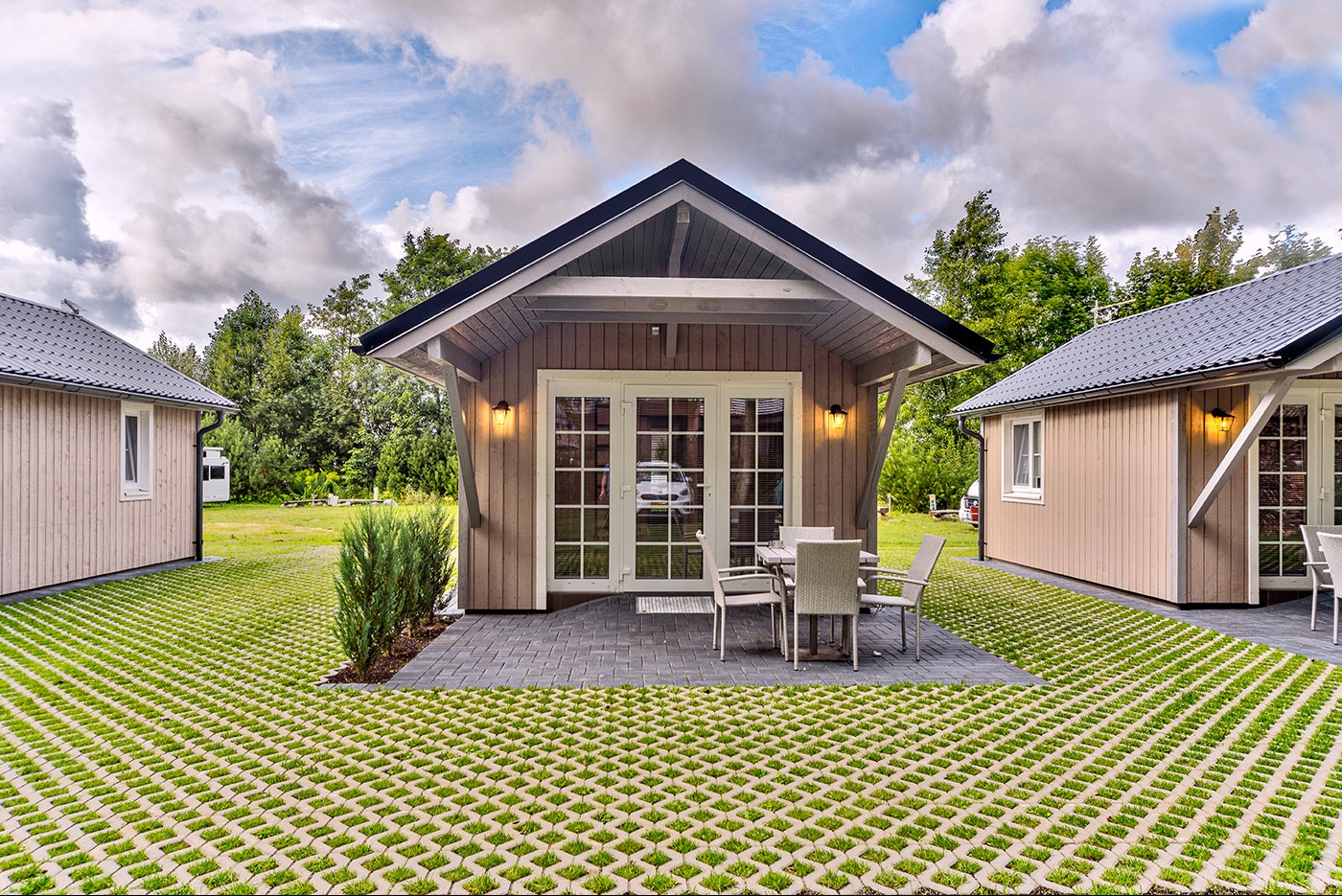 Holzhaus mit Veranda und Sitzplatz, davor ein gepflegter Rasen mit Zierbeeten, daneben weitere Ferienhäuser im Campingbereich.