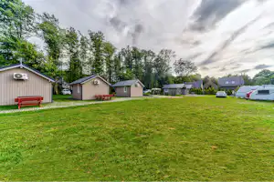 Grüner Campingplatz mit Rasenfläche, mehreren kleinen Ferienhäusern und Wohnwagen sowie Bank und Bäumen am Rand.