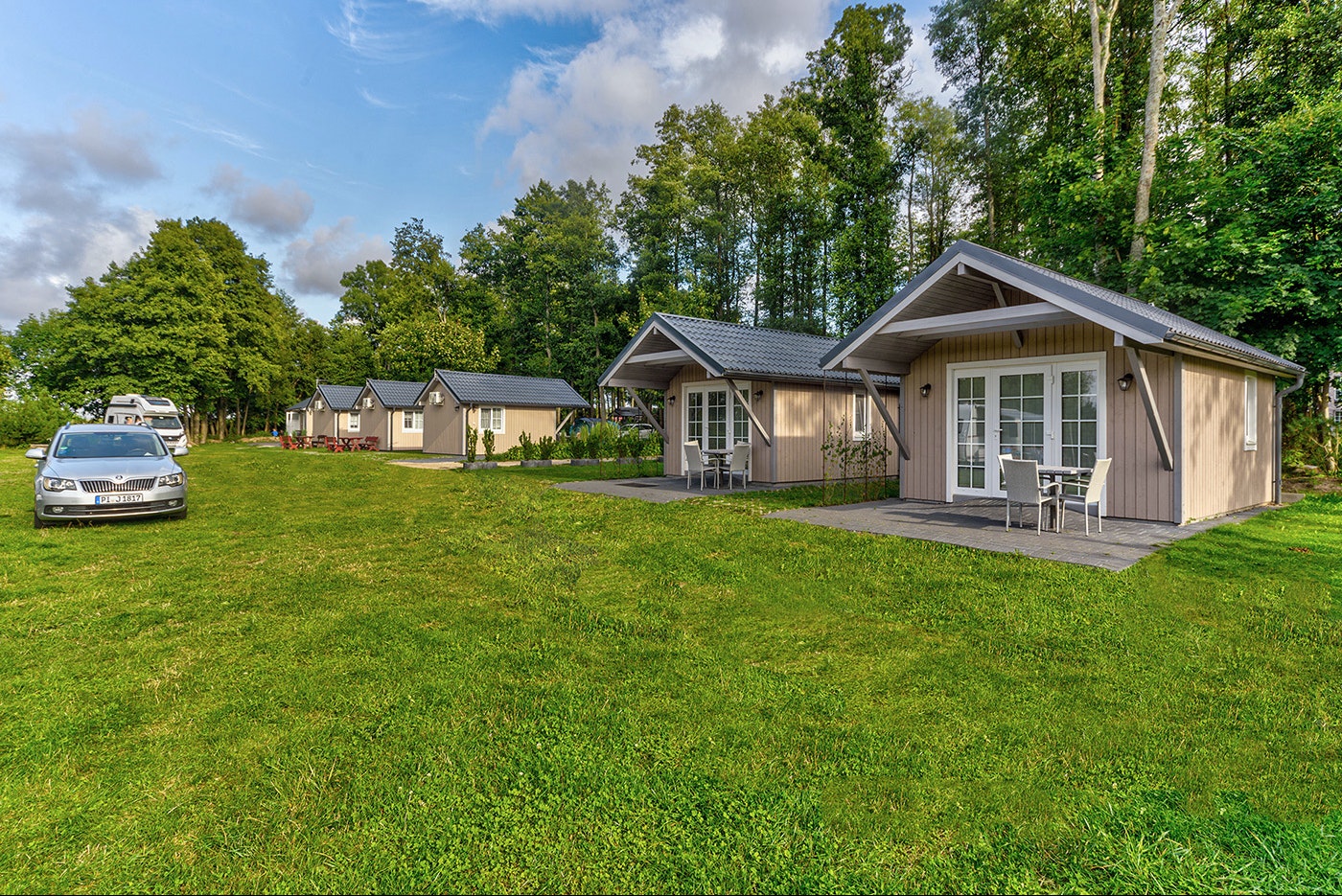 Reihen von Bungalows auf einem grünen Campingplatz, mit Terrasse und Rasenfläche, im Hintergrund Bäume und ein Auto.