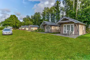 Reihen von Bungalows auf einem grünen Campingplatz, mit Terrasse und Rasenfläche, im Hintergrund Bäume und ein Auto.