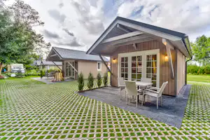 Holzhaus mit Terrasse und Sitzgruppe, gepflasterter Bereich und gepflegter Rasen im Campinggarten, Bäume im Hintergrund.