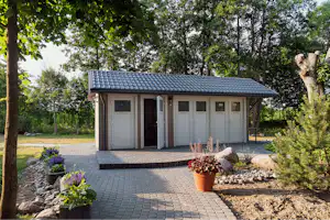 Kleines Sanitärgebäude mit Schiebetüren und Ziegeldach, gepflasterter Vorplatz, Blumen in Töpfen und Beeten, umgeben von Bäumen.