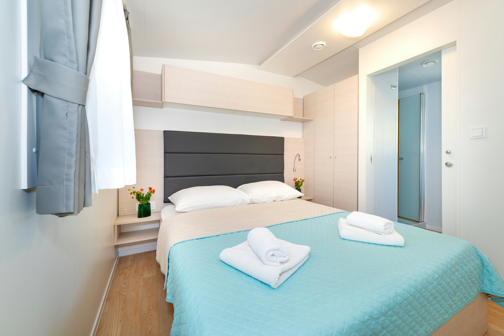 Modernes Camping-Schlafzimmer mit Doppelbett, blauer Bettdecke und gefalteten Handtüchern, daneben Vorhang und Nachttisch.