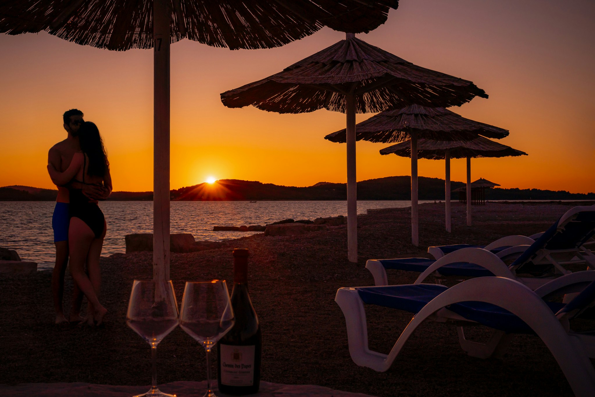 Sonnenuntergang am Strand: Liegen und Sonnenschirme, daneben Gläser und eine Flasche auf dem Campingplatz.