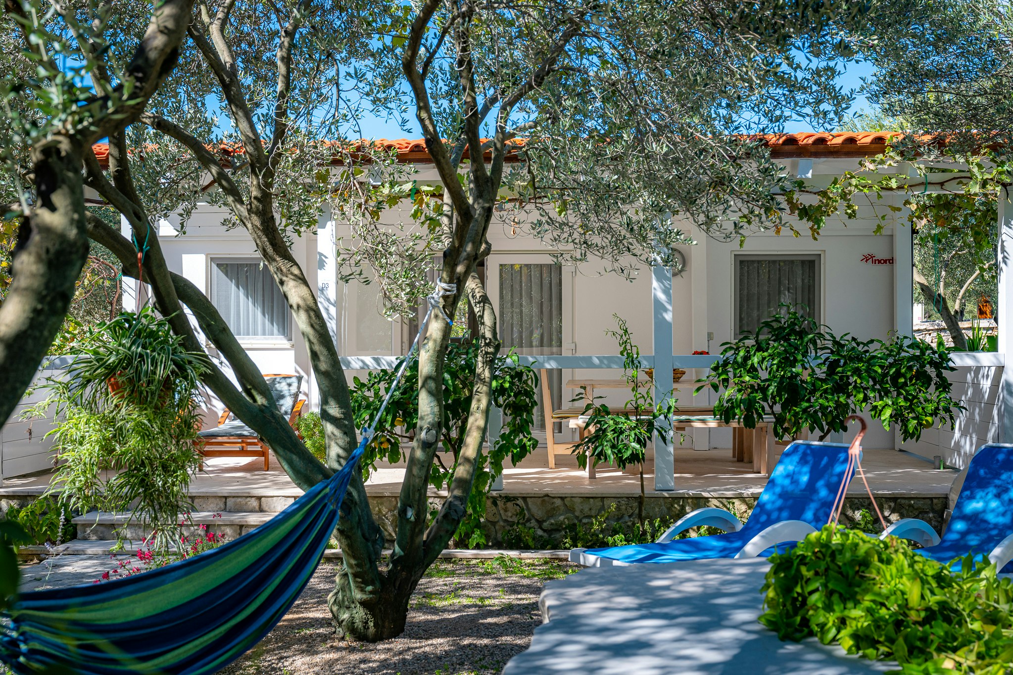 Hängematte im Garten zwischen Bäumen vor einem weißen Ferienhaus mit Terrasse und Sitzbank, gepflasterter Weg im Vordergrund.
