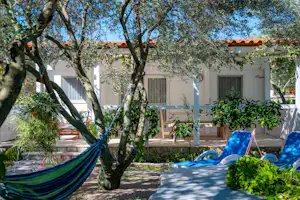 Hängematte im Garten zwischen Bäumen vor einem weißen Ferienhaus mit Terrasse und Sitzbank, gepflasterter Weg im Vordergrund.