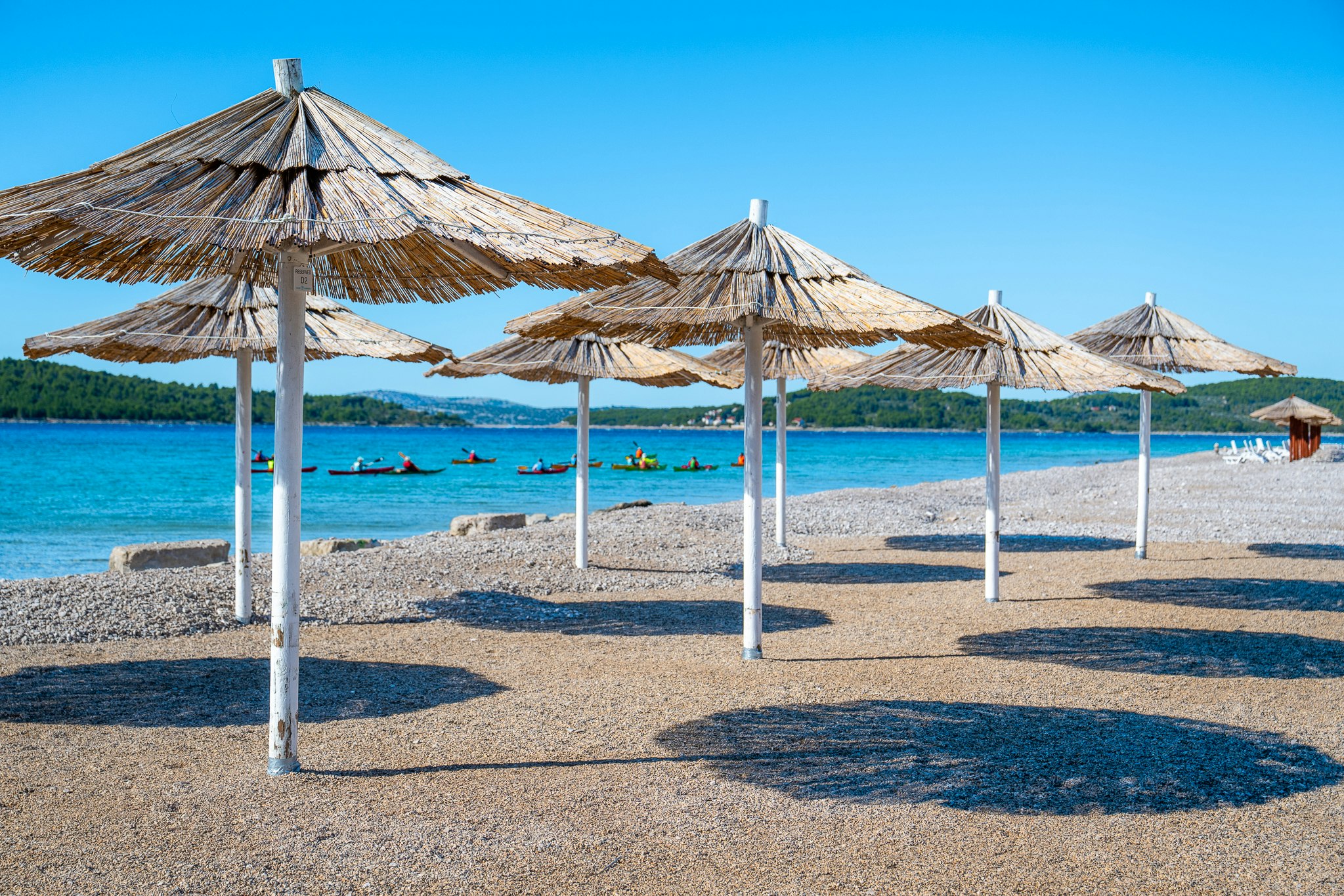 Strand mit mehreren Sonnenschirmen auf Kies, dahinter das blaue Meer und einzelne Boote im Wasser.