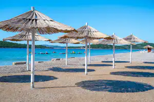Strand mit mehreren Sonnenschirmen auf Kies, dahinter das blaue Meer und einzelne Boote im Wasser.