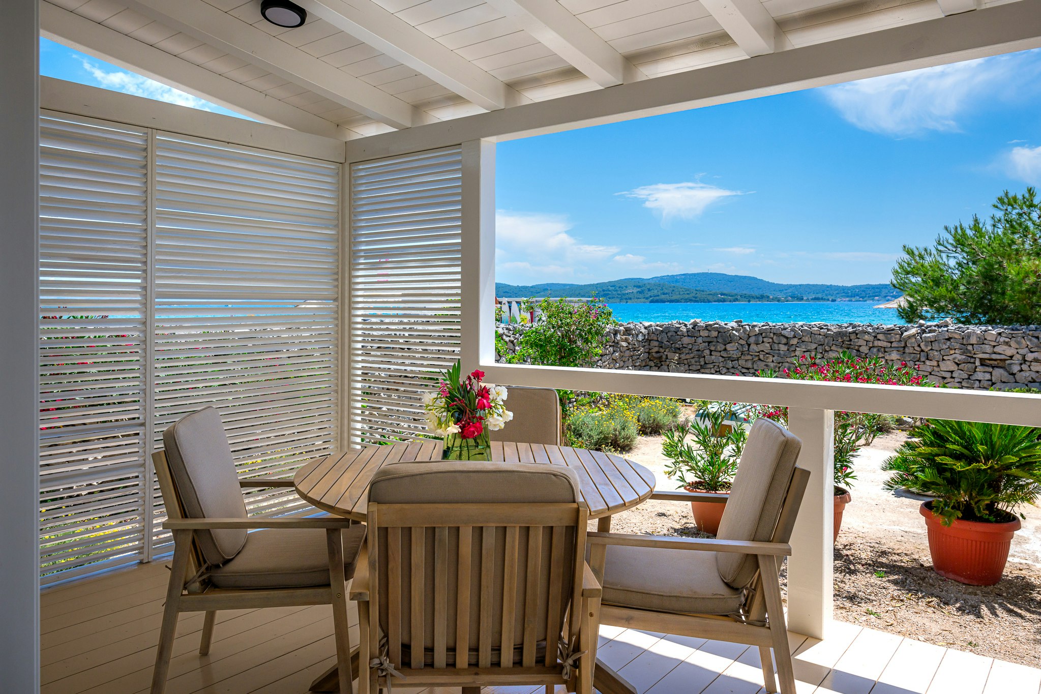 Terrasse mit Holztisch und Stühlen, Blick auf das Meer und die Küste, Steinmauer und Pflanzen im Vordergrund.