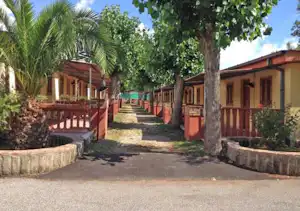 Weg zwischen Bungalows mit Veranda, Holzgeländern und Sitzbereichen, umgeben von Palmen und Bäumen.