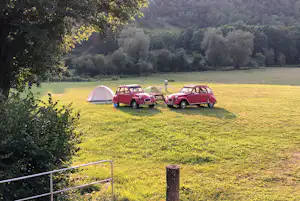 Zelt und zwei rote Oldtimer-Kleinwagen auf einer Campingwiese, umgeben von Bäumen und Hügeln im Hintergrund.