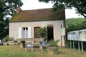 Ferienhaus mit rotem Ziegeldach, weißen Fenstern und Eingangstür, davor Garten mit Blumen und zwei Metallstühlen.