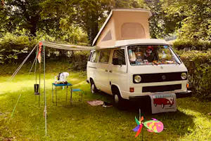 Weißes Wohnmobil mit aufgestelltem Zelt auf dem Campingplatz, daneben Tisch und Stühle im Grünen.
