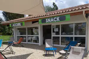 Snack-Bereich am Campingplatz mit Terrasse, blauen Tischen und Stühlen unter Sonnenschirm, neben dem Gebäude mit Schaufenstern.