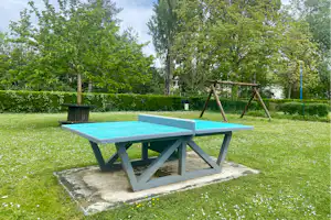Blauer Camping-Tisch auf Betonplatte im grünen Garten, mit Hecke, Bäumen und Spielschaukel im Hintergrund.