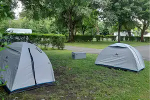 Zwei Campingzelte auf einer grünen Wiese zwischen Bäumen, mit einem kleinen Holzkasten und Wegen am Rand.