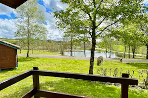 Blick vom Holzgeländer auf den Campingplatz mit See, grüner Wiese, Bäumen und einem Weg zwischen den Stellplätzen.