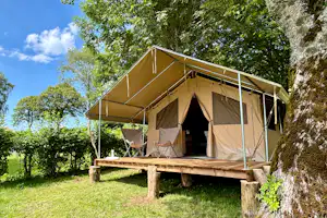 Glamping-Zelt mit Holzterrasse, zwei Klappstühlen und kleinem Tisch im Grünen, umgeben von Bäumen und Sträuchern.