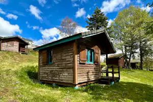 Holzhütte mit Veranda, braunen Türen und grünen Fensterrahmen auf einer Wiese, umgeben von Bäumen und weiteren Hütten.