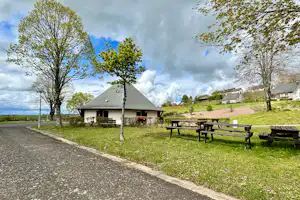 Campinghaus mit Satteldach, Picknicktischen und Bäumen am Weg, im Hintergrund weitere Gebäude auf grüner Wiese.