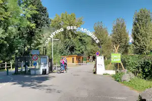 Eingang zum Campingplatz Des Adourbes mit Schild, Schranke und breitem Weg, umgeben von Bäumen und Hecken.
