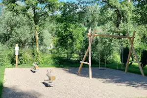 Spielplatz auf dem Campingplatz mit Sandfläche, Schaukelgestell und Bäumen, im Hintergrund grüne Hügel und Berge.