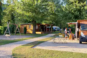Parkplatz mit orangefarbenem Wohnmobil und Kiesweg, daneben Wohnwagen und Spielplatz im Grünen mit Bäumen.
