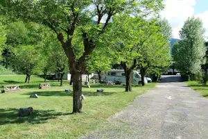 Grüner Campingplatz mit Kiesweg, Bäumen und Rasenflächen; einzelne Liegen und Sitzgelegenheiten im Schatten.