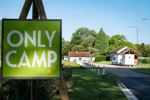 Schild „ONLY CAMP!“ am Campingplatz, daneben Wohnwagen und Gebäude am Weg, Bäume und Rasen im Hintergrund.