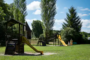 Spielplatz am Campingplatz mit Rutsche, Holzunterstand und Sitzbank auf grünem Rasen, umgeben von Bäumen und Wegen.