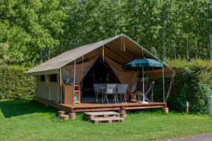 Großes Campingzelt auf Holzpodest mit Vorzelt, Tisch und Stühlen, umgeben von Rasen und Bäumen im Grünen.
