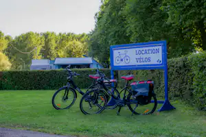 Blaues Schild „Location Vélos“ mit Fahrrädern vor Hecken und Wohnwagen auf dem Campingplatz.
