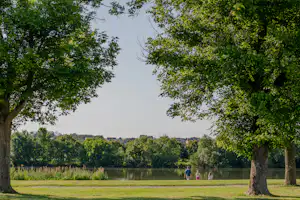 Blick auf einen See im Campingpark, umrahmt von Bäumen und Gras, mit Spaziergängern am Ufer.
