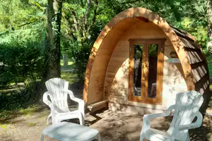 Holzhütte mit Eingangstür und zwei weißen Kunststoffstühlen auf dem Stellplatz im Grünen, umgeben von Bäumen.