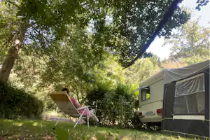 Wohnwagen im Campinggarten mit Markise, Liegestuhl und viel Grün unter hohen Bäumen.