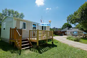 Mobilheim mit Holzterrasse, Stufen und Markise, im Hintergrund weitere Gebäude und Flaggen vor blauem Himmel.