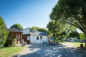 Campinggebäude mit Eingang, Infotafeln und Sitzbank vor einer Wiese, daneben Zelte und Fahrräder unter großen Bäumen.