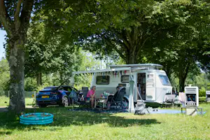 Wohnmobil mit Markise und Campingstühlen, daneben Auto, auf einer Wiese unter großen Bäumen.