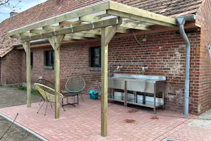 Grillplatz unter Holzüberdachung an einer Ziegelwand mit Edelstahl-Grill, Wasserhähnen und Sitzgelegenheit auf Pflastersteinen.
