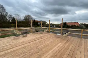 Holzterrasse mit Sitzgruppe aus Korbsesseln und kleinen Tischen, Blick auf Bäume und Häuser bei bewölktem Himmel.