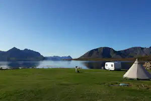 Campingzelt auf einer Wiese am See, im Hintergrund Wohnmobil und Berge, ruhiges Wasser und Uferlinie.