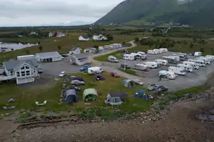 Campingplatz im Tal mit Wohnwagen und Zelten, mehreren Fahrzeugen und Wohnhäusern am Hang, umgeben von Bergen.