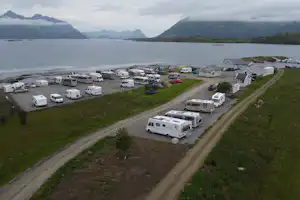 Wohnmobile und Campingplätze entlang eines breiten Weges, am Ufer mit Wasser und Bergen im Hintergrund.