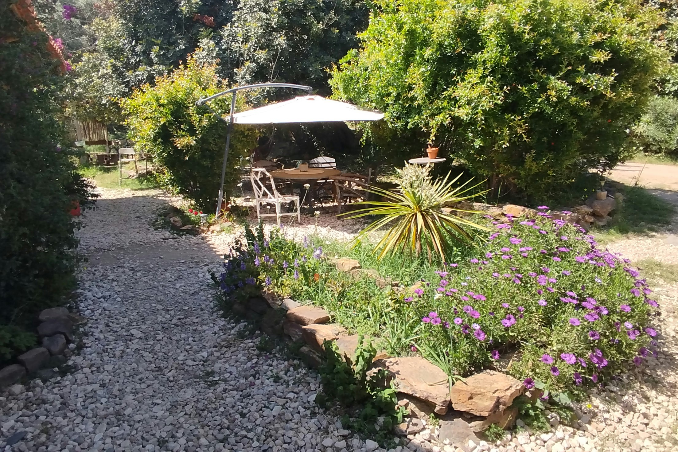 Gartenweg mit Kiesfläche, Blumenbeet und Sitzbereich unter einem Sonnenschirm zwischen Bäumen und Hecken.