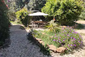 Gartenweg mit Kiesfläche, Blumenbeet und Sitzbereich unter einem Sonnenschirm zwischen Bäumen und Hecken.