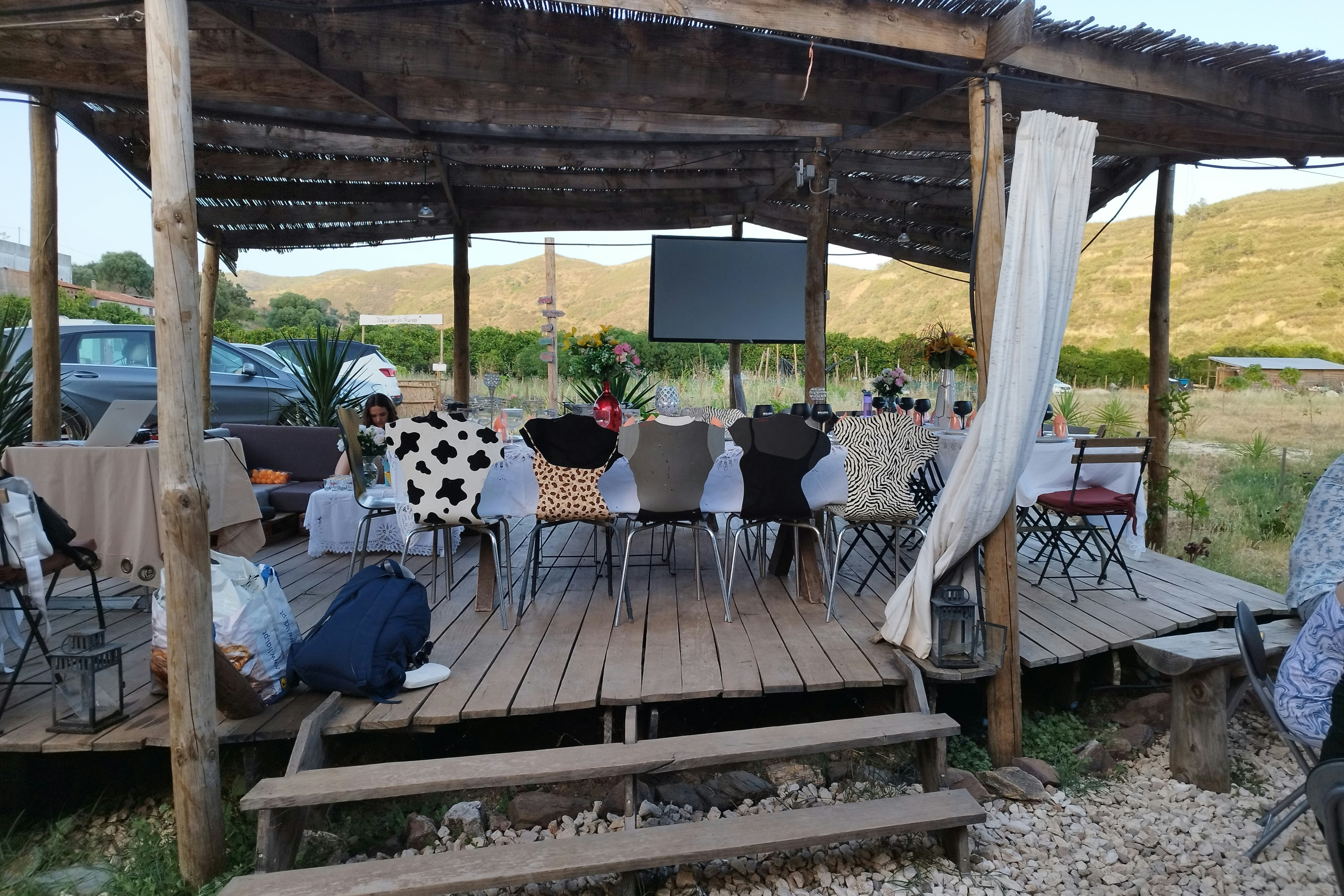 Überdachte Holzterrasse mit Sitzgruppe und Tisch, Deko-Kissen und Vorhängen, Blick auf Hügel und Zeltbereich im Hintergrund.