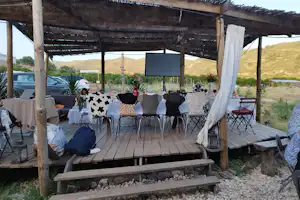 Überdachte Holzterrasse mit Sitzgruppe und Tisch, Deko-Kissen und Vorhängen, Blick auf Hügel und Zeltbereich im Hintergrund.