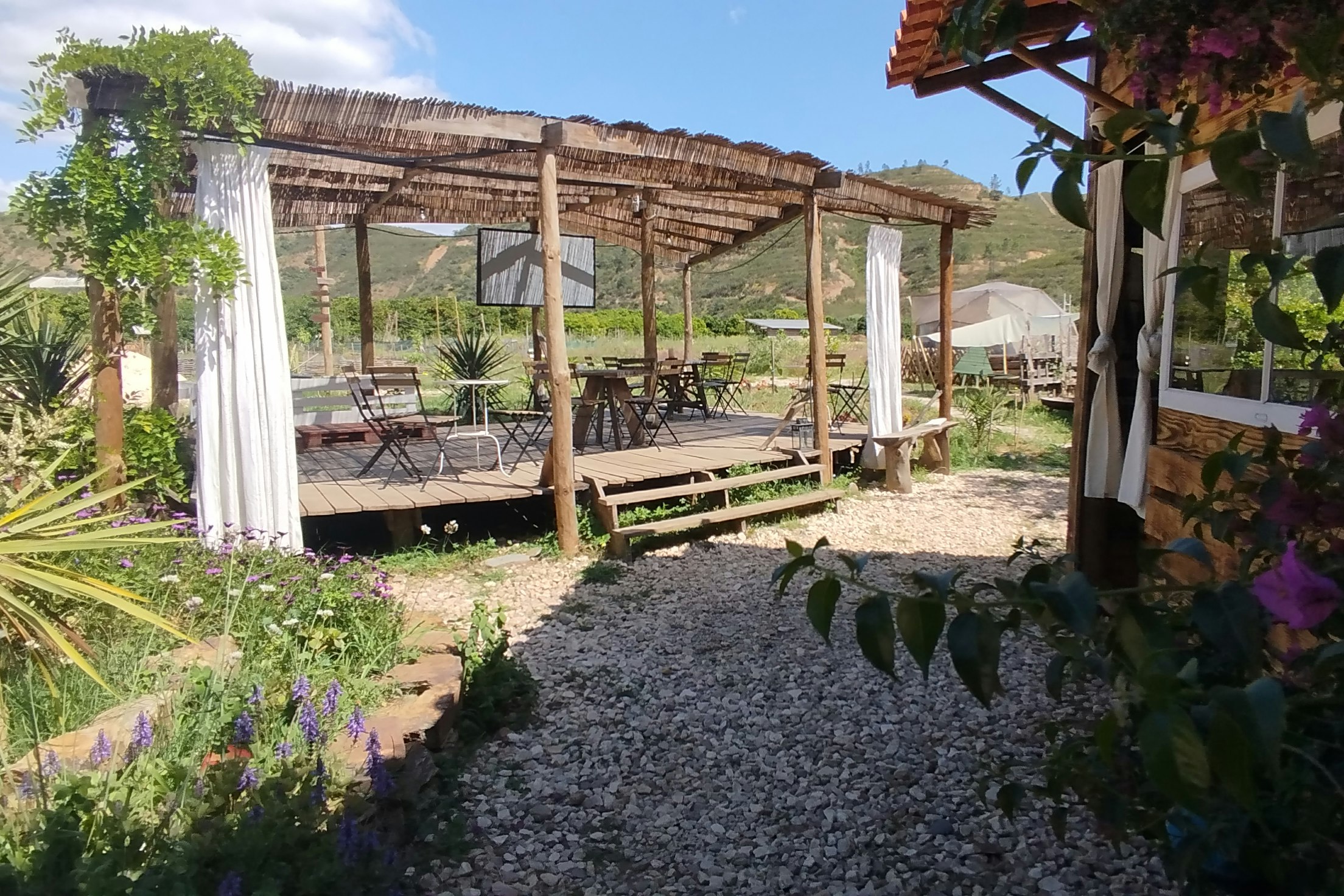 Holzpergola mit Sitzbereich auf einem Campingplatz, Kiesweg und Pflanzen im Vordergrund, Berge im Hintergrund.