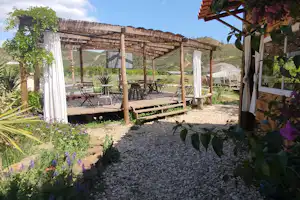 Holzpergola mit Sitzbereich auf einem Campingplatz, Kiesweg und Pflanzen im Vordergrund, Berge im Hintergrund.